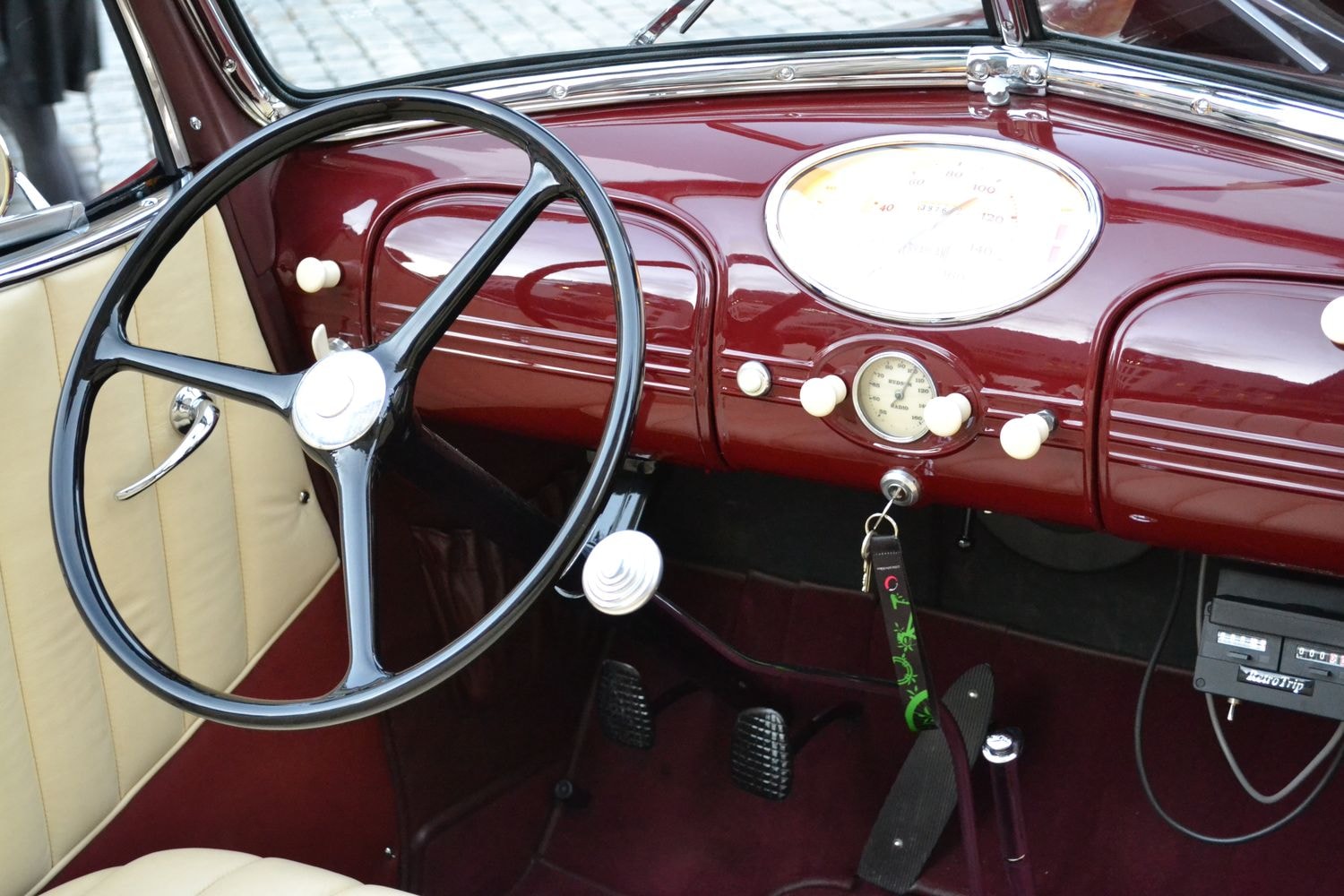 Innenansicht eines Oldtimers mit einem cremefarbenen Ledersitz und einem dunkelroten Armaturenbrett. Das Lenkrad ist schwarz und hat ein klassisches Design. Auf dem Armaturenbrett befinden sich mehrere Knöpfe und Anzeigen, darunter ein großer Tacho in der Mitte und eine kleinere Uhr darunter. Ein Schlüsselbund hängt an einem grünen Band unterhalb des Armaturenbretts.