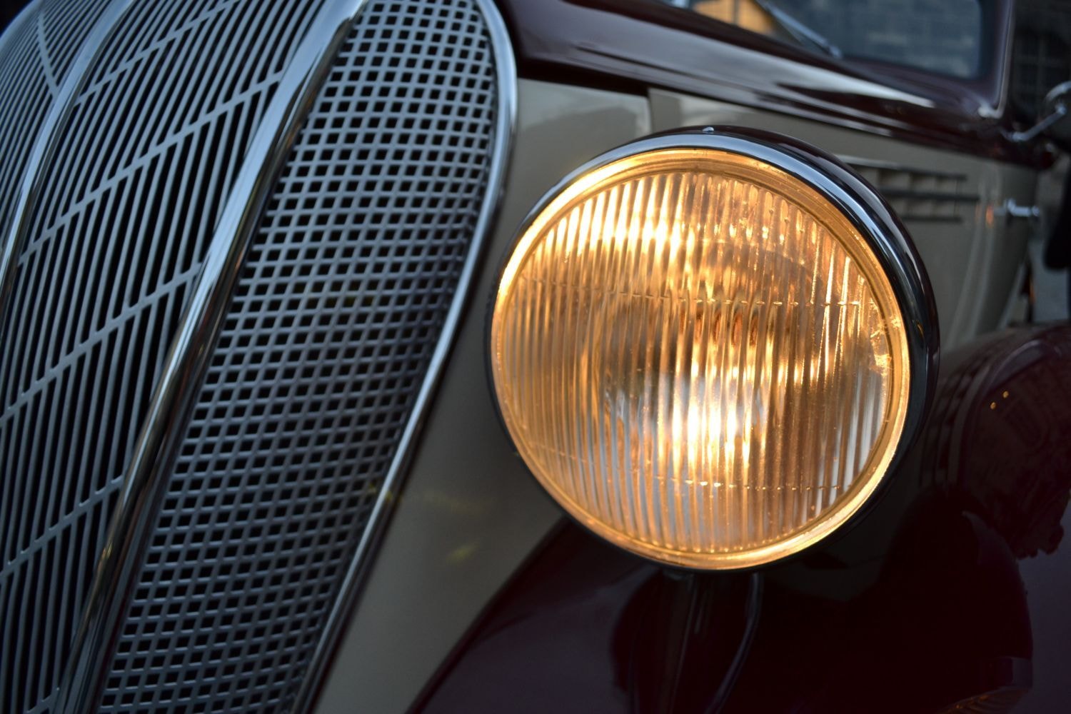 "Eine Nahaufnahme des Vorderteils eines Oldtimers, fokussiert auf den Scheinwerfer und den Kühlergrill. Der Scheinwerfer ist beleuchtet und strahlt ein warmes gelbes Licht aus. Der Kühlergrill hat ein detailliertes Maschenmuster.