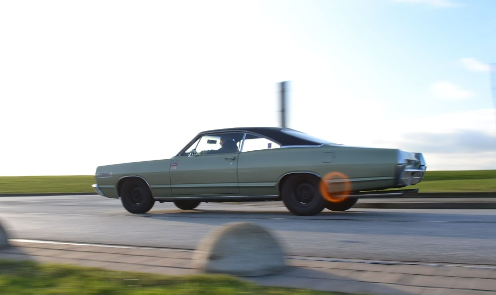 Ein grünes, klassisches Auto fährt auf einer Straße entlang. Im Hintergrund sind eine grüne Wiese und ein heller Himmel zu sehen. Ein Lichtreflex ist an der hinteren Seite des Autos sichtbar.