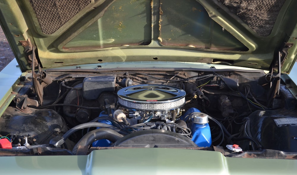 Der Motorraum eines Autos mit geöffneter Motorhaube zeigt einen V8-Motor mit verchromter Luftfilterabdeckung. Der Motorblock und die Ventildeckel sind blau lackiert. Die Karosserie des Autos ist grün.