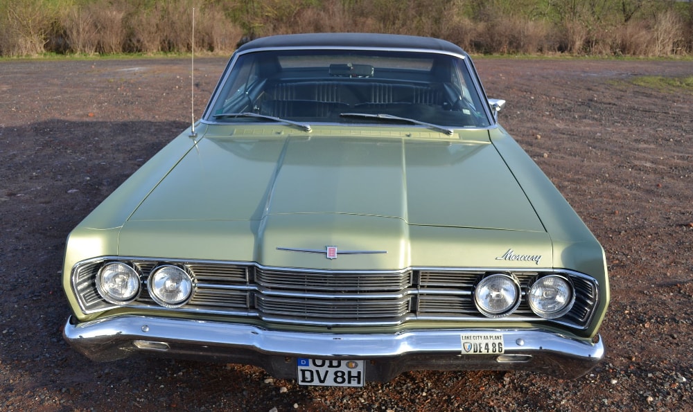 Die Vorderansicht eines hellgrünen Mercury Monterey mit Chromdetails, runden Doppelscheinwerfern und den Nummernschildern "DUB DV 8H" und "CM 4298" aus Michigan. Das Auto steht auf einer Kiesfläche mit Vegetation im Hintergrund.