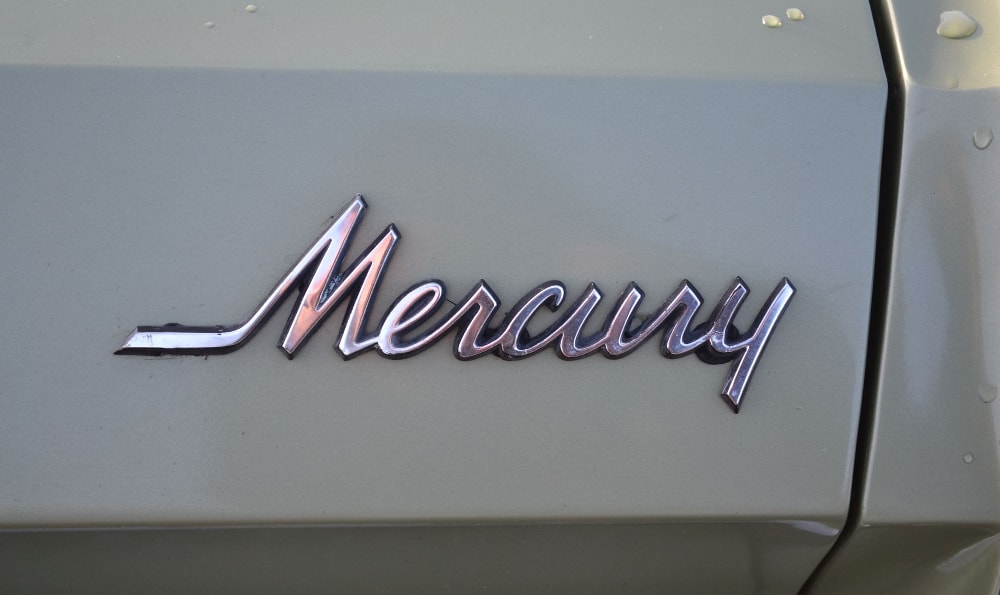 Das Bild zeigt das Emblem eines Mercury-Fahrzeugs. Das Emblem ist in einer kursiven Schrift und metallisch, reflektiert das Licht. Der Hintergrund scheint die Karosserie des Autos zu sein, die in einer hellen Farbe lackiert ist.