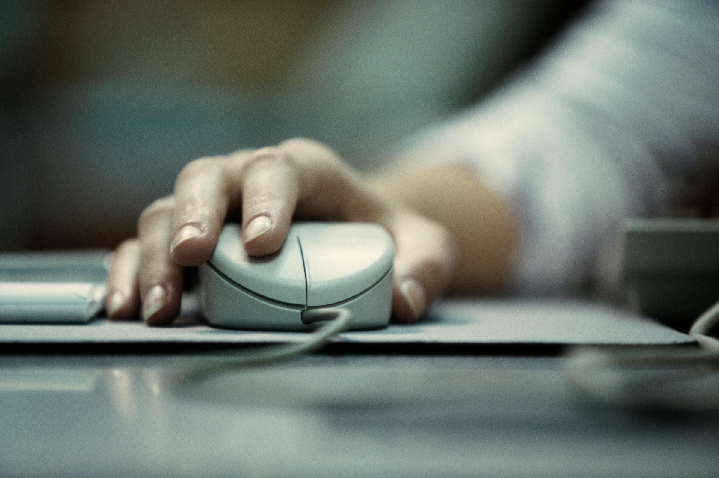 Eine Nahaufnahme einer Hand, die auf einer Computermaus ruht. Die Hand ist in Position, um die linke Maustaste zu klicken, und ein Kabel ist an der Vorderseite der Maus sichtbar. 