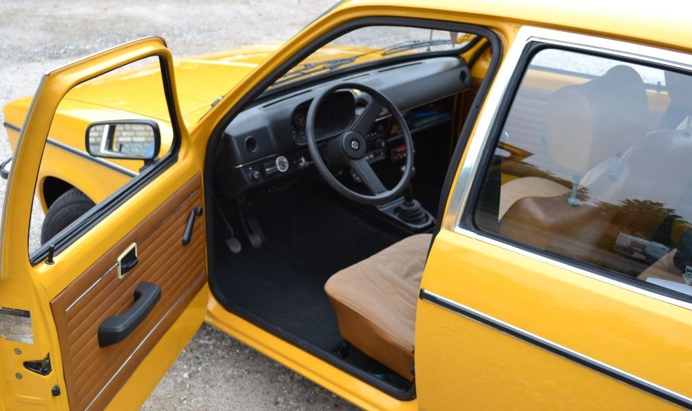 Innenraum eines gelben Autos mit geöffneter Fahrertür, brauner Polsterung und Schalthebel.