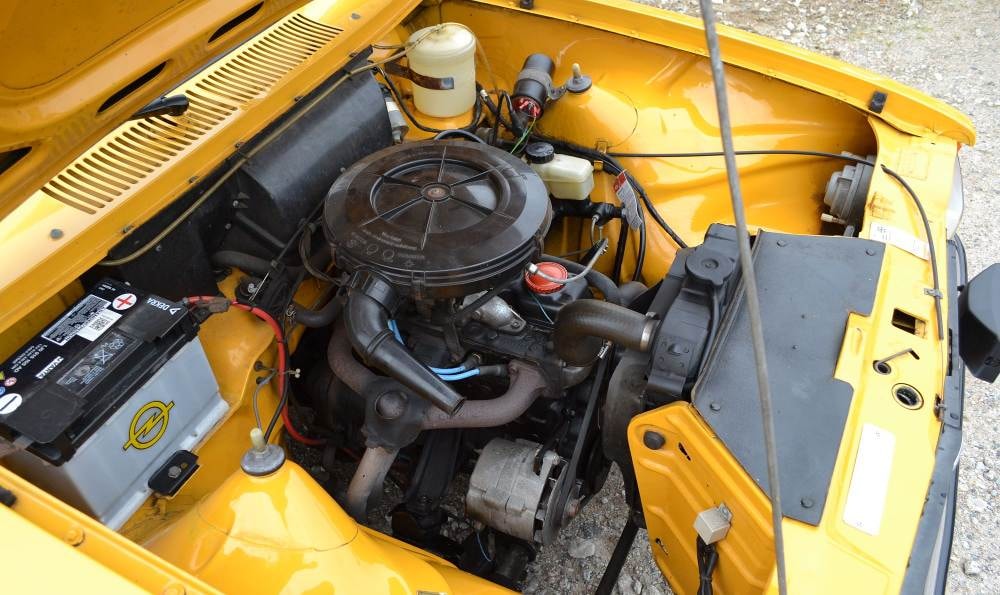 Motorraum eines gelben Autos mit zentralem Motor, Batterie links und Kühlmittelbehälter oben.