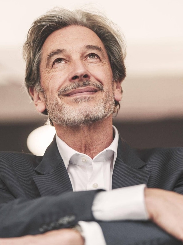 Person im dunklen Anzug mit weißem Hemd und verschränkten Armen, Gesicht unscharf.