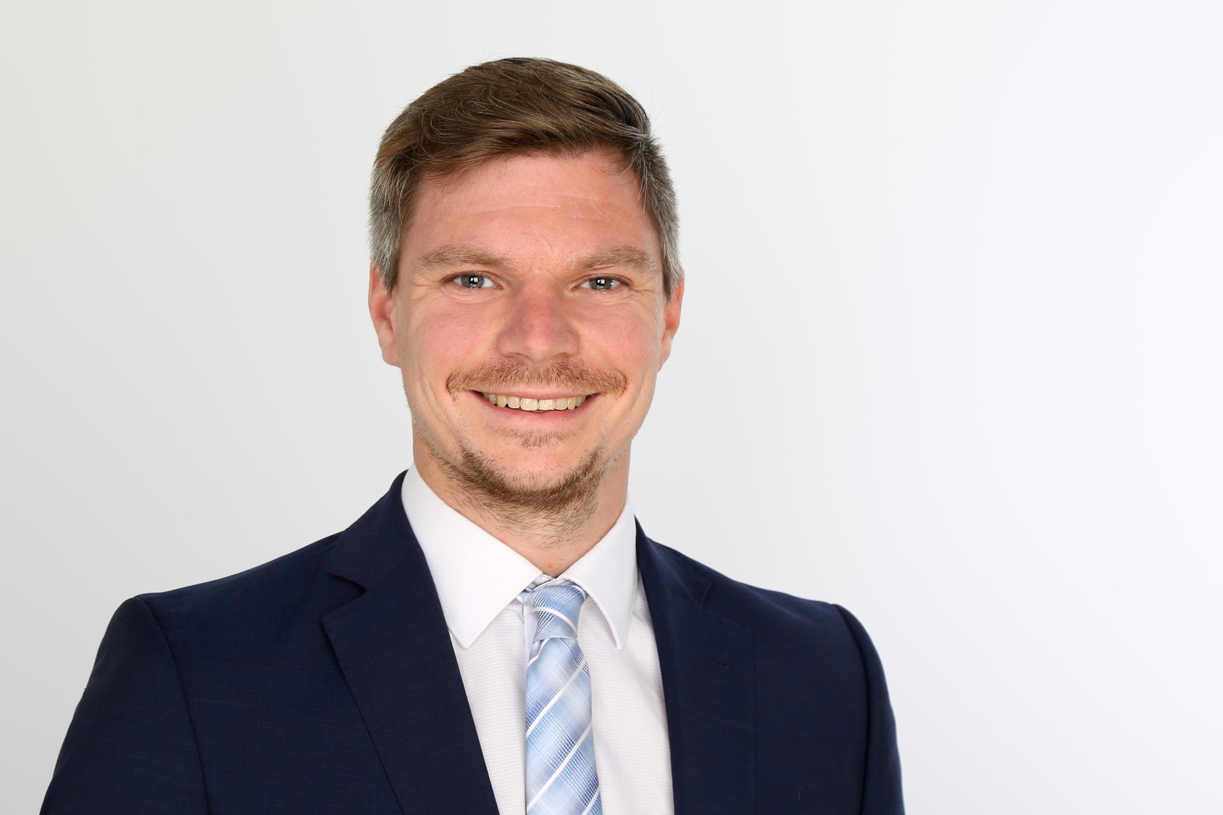 Tobias Knoll trägt einen dunklen Anzug und weißem Hemd vor weißem Hintergrund.