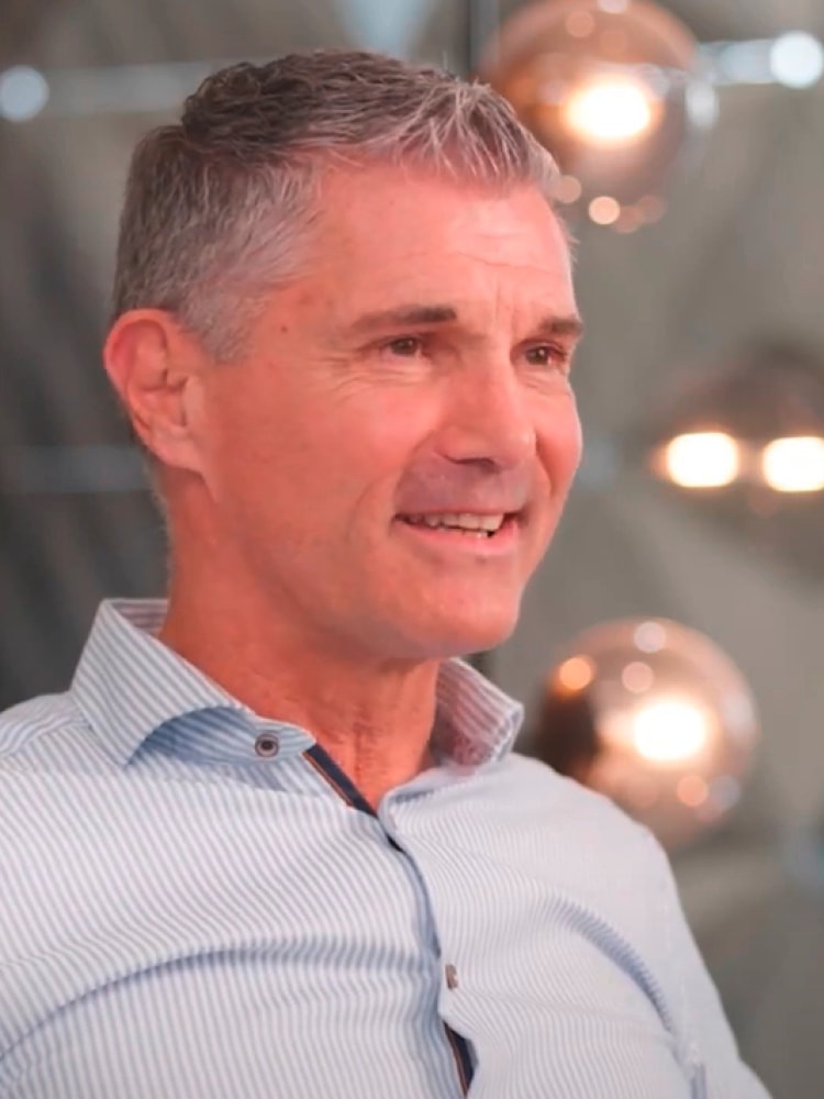 as Bild zeigt einen Mann mittleren Alters mit grauen Haaren, der ein gestreiftes Hemd trägt. m Hintergrund sind verschwommene Lichter zu sehen, die eine warme und gemütliche Atmosphäre schaffen