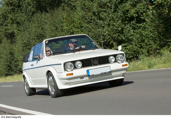 Weißes Cabriolet mit zwei Personen fährt auf einer Straße, im Hintergrund Bäume; blaues Nummernschild sichtbar.