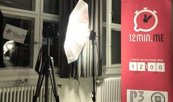 Veranstaltungsaufbau mit rotem Banner von 12min.me, einem Stativ und einer Beleuchtungseinrichtung in einem Raum mit großen Fenstern