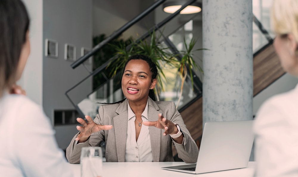 Eine Frau in einem beigen Anzug gestikuliert ausdrucksstark während einer Besprechung. Sie sitzt an einem Tisch mit einem Laptop und unterhält sich mit zwei Personen, was eine Atmosphäre der Zusammenarbeit vermittelt.