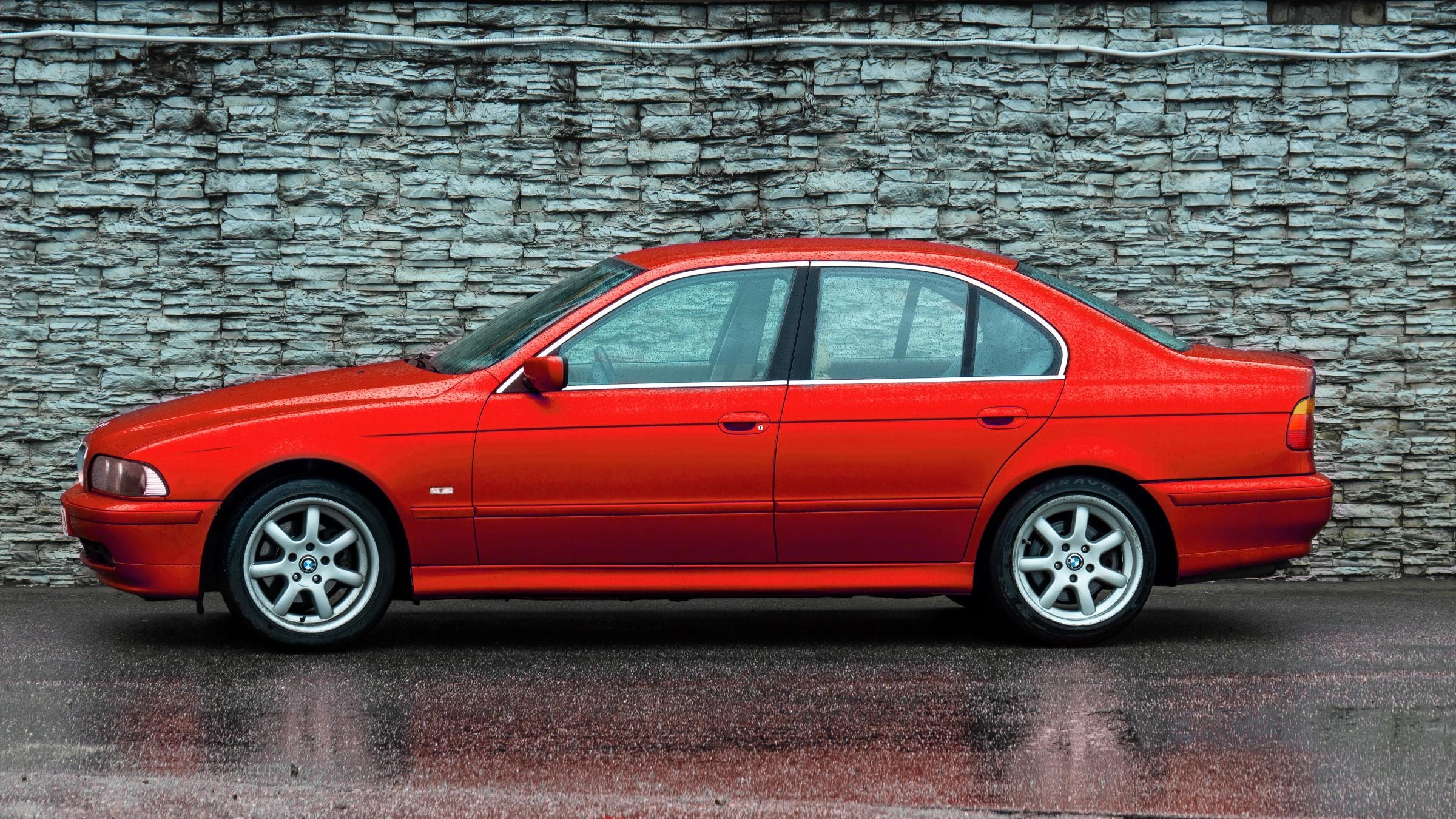 Ein roter BMW E39 Limousine parkt bei Regen vor einer grauen Ziegelwand.