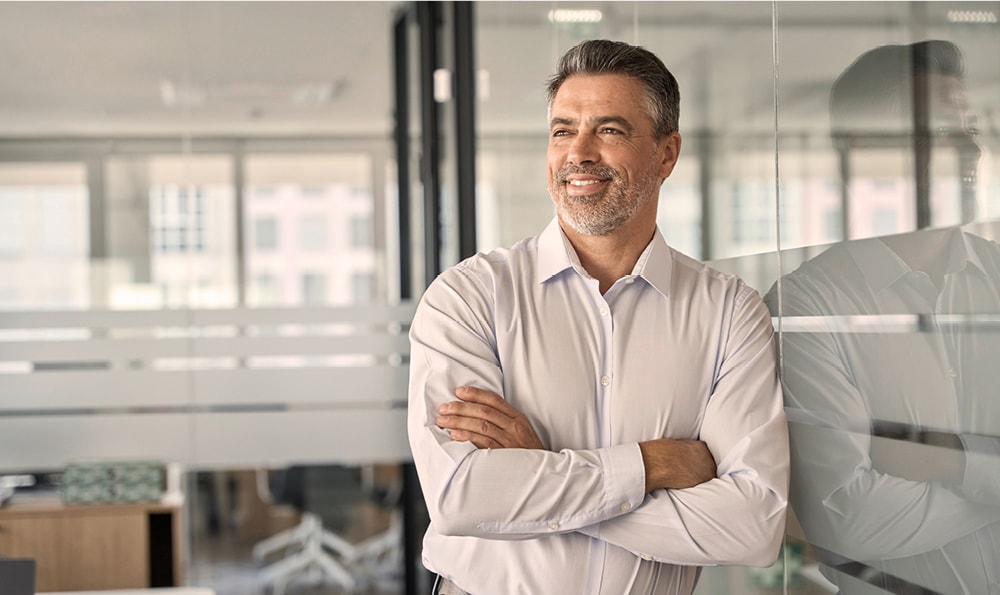 Ein Mann mit grauen Haaren und Bart, der ein helles Hemd trägt, steht lächelnd mit verschränkten Armen in einem modernen Büro.