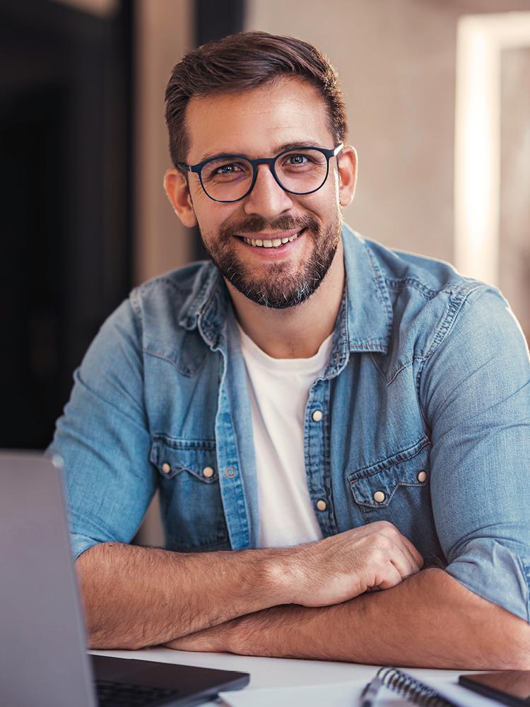 Ein junger Mann mit Brille, Bart und in Jeanshemd hat die Hände vor seinem Laptop verschränkt und blickt lächelnd in die Kamera