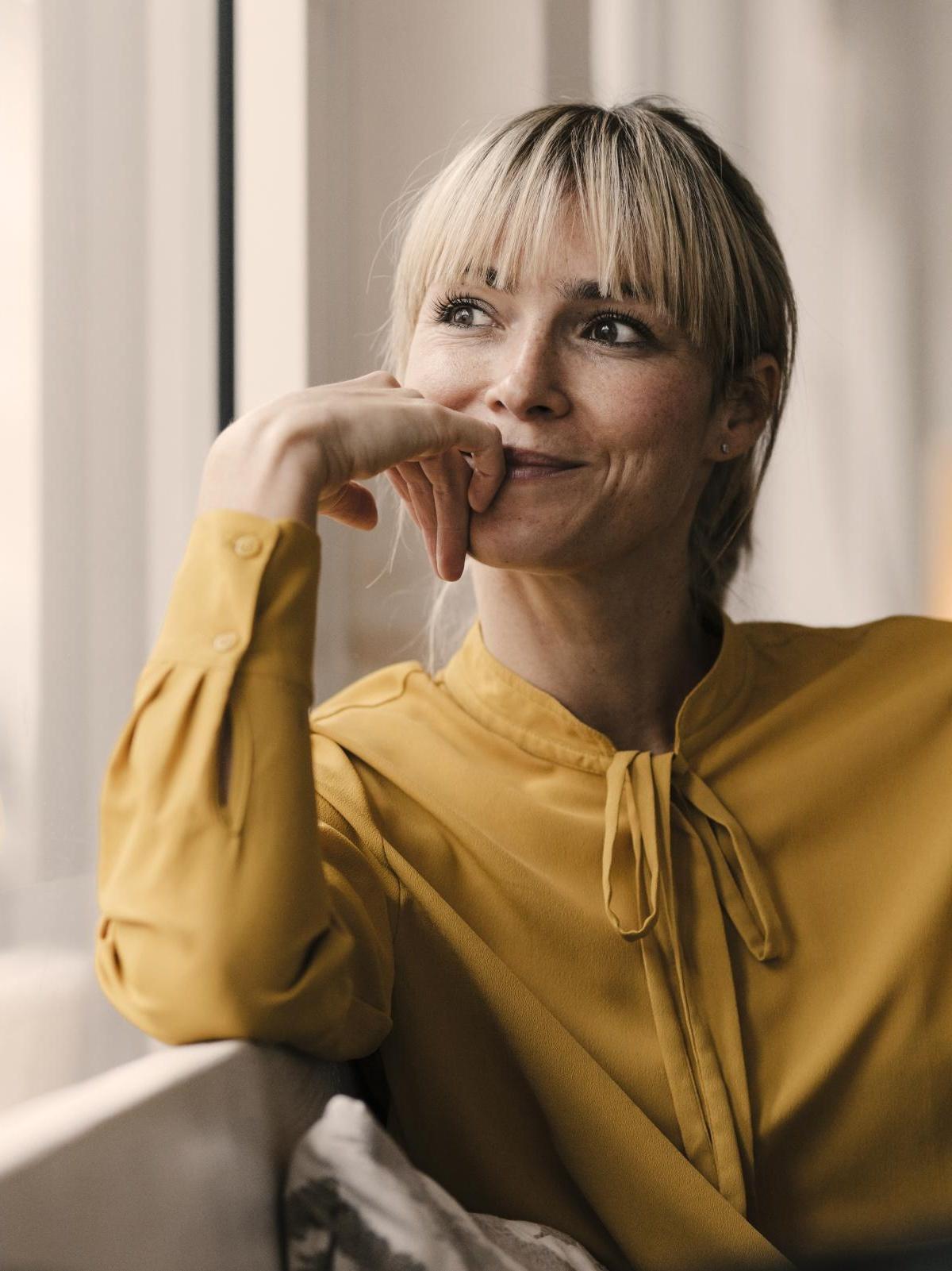 Junge blonde Businessfrau in gelber Bluse sitzt am Fenster und schaut nach draußen.