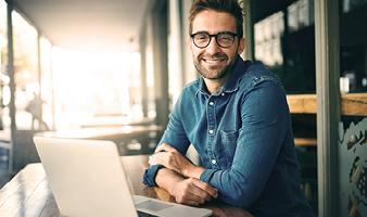 Ein Mann in Hemd und mit Brille sitzt vor seinem Laptop und lächelt in die Kamera