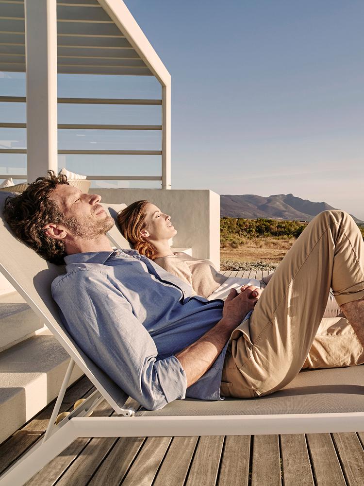 Eine Frau und ein Mann sonnen sich auf Liegen auf einer Terrasse, im Hintergrund sich Berge zu sehen