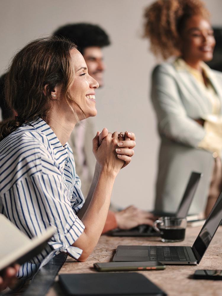 Eine junge Frau mit gestreiftem Hemd sitzt vor dem Laptop und lächelt nach rechts. Im Hintergrund stehen weitere junge Menschen, die gemeinsam arbeiten