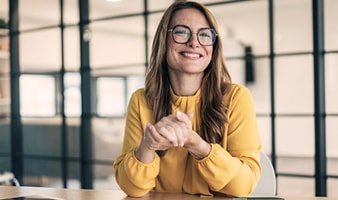 Eine Frau in gelber Bluse sitzt an einem Schreibtisch in einem modernen Büro und gestikuliert mit den Händen.
