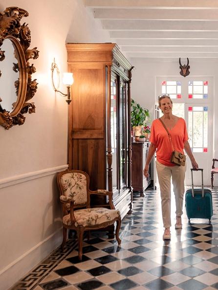 Eine lächelnde Frau in einem orangefarbenen Oberteil zieht einen blaugrünen Koffer durch einen hellen, eleganten Flur mit kariertem Boden, einem Vintage-Sessel und einem dekorativen Spiegel.