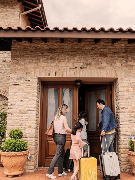Eine vierköpfige Familie betritt mit ihrem Gepäck ein rustikales Steinhaus. Die Stimmung ist einladend und warm, die Reisenden sind bereit, ihren Aufenthalt zu beginnen.ox