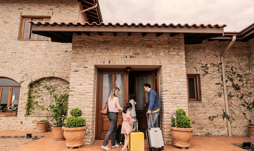 Familie bei der Ankunft in einem Steinhaus. Ein Mann öffnet die Eingangstür, während eine Frau und zwei Kinder mit Gepäck daneben stehen. Die Szene wirkt warm und einladend.