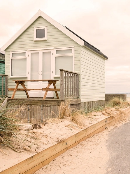 Eine hellgrüne Strandhütte mit einer kleinen Veranda und einem Picknicktisch liegt in den Sanddünen unter einem wolkenverhangenen Himmel, der eine ruhige Küstenatmosphäre schafft.