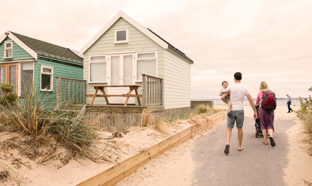 Eine Familie geht auf einem Sandweg zwischen Strandhütten spazieren, wobei ein Mann ein Kind trägt und eine Frau einen Kinderwagen schiebt. Die Szene wirkt warm und entspannend.