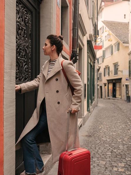 Eine Frau mit einem roten Koffer betritt eine Tür in einer engen italienischen Straße. Sie trägt einen beigen Mantel und Jeans, die an Reisen und Entdeckungen erinnern.