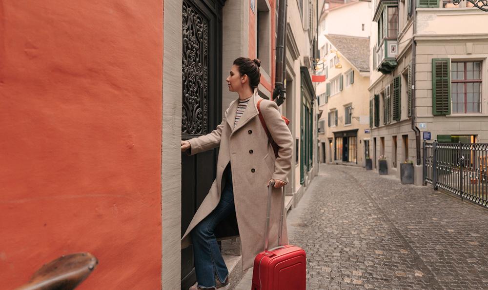 Eine Frau in einem beigen Mantel und mit einem roten Koffer in der Hand betritt eine Tür in einer engen, kopfsteingepflasterten europäischen Straße. Die Szene wirkt heiter und entdeckend.