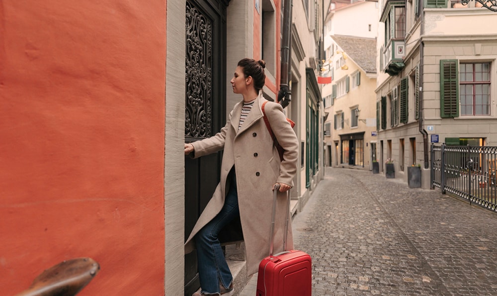 Eine Frau in einem beigen Mantel und mit einem roten Koffer in der Hand betritt eine Tür in einer engen, kopfsteingepflasterten europäischen Straße. Die Szene wirkt heiter und entdeckend.