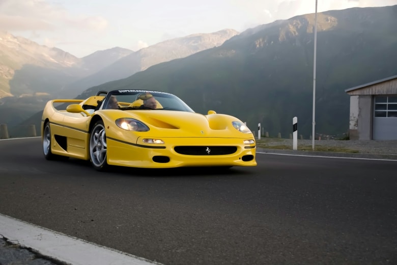 Gelber Ferrari F50 fährt auf einer Bergstraße mit Bergszenario im Hintergrund.