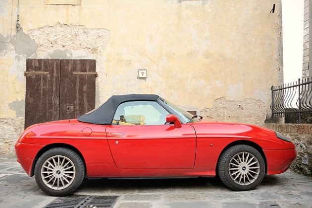 Roter Fiat Barchetta parkt seitlich vor einer sandfarbenen Wand. Das Verdeck ist geschlossen.