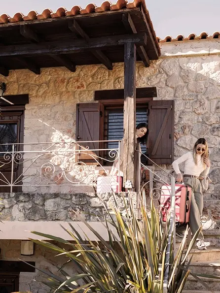 Zwei Frauen steigen mit rosafarbenem Gepäck eine Steintreppe an einem rustikalen Steinhaus hinunter. Sie wirken entspannt und glücklich und sonnen sich im warmen Sonnenlicht.