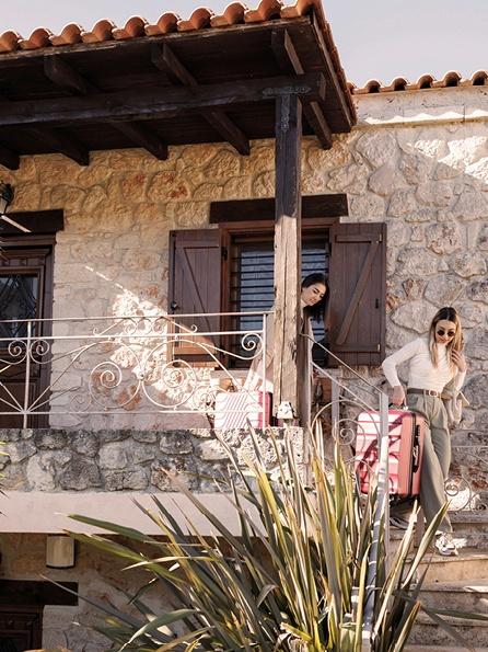 Zwei Frauen steigen mit rosafarbenem Gepäck eine Steintreppe an einem rustikalen Steinhaus hinunter. Sie wirken entspannt und glücklich und sonnen sich im warmen Sonnenlicht.