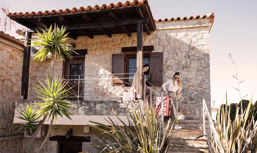 Zwei Personen stehen auf den Stufen eines rustikalen Steinhauses, eine trägt einen Koffer. Palmen flankieren die Treppe unter einem klaren Himmel.