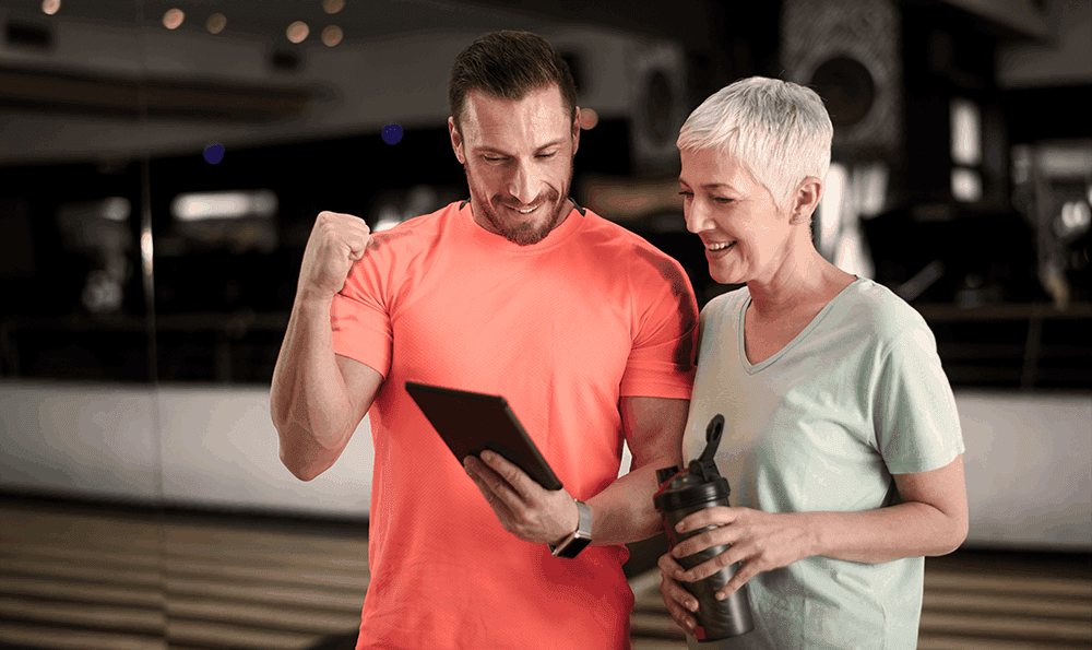 fiEin Mann in einem leuchtend orangefarbenen Shirt und eine Frau in einem hellen Shirt stehen in einem Fitnessstudio und lächeln auf ein Tablet. Der Mann feiert mit einer Faustbewegung und drückt damit seine Begeisterung aus.