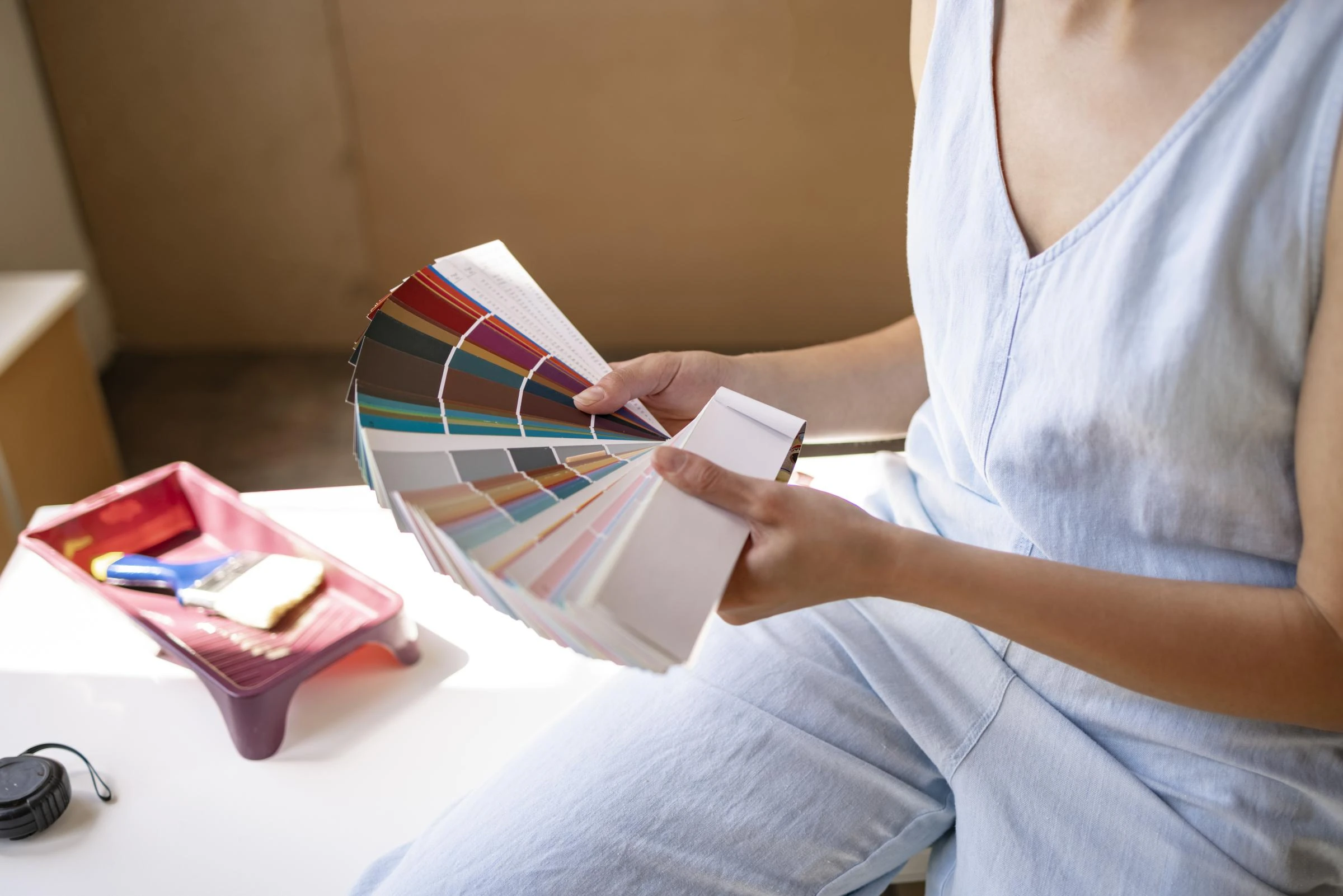 Eine Person in blauer Kleidung hält einen Fächer mit Farbmusterkarten in der Hand, was auf Innenarchitektur oder Malerei hindeutet. In der Nähe deutet ein Tablett mit Pinseln und Klebeband auf Vorbereitungsarbeiten hin.
