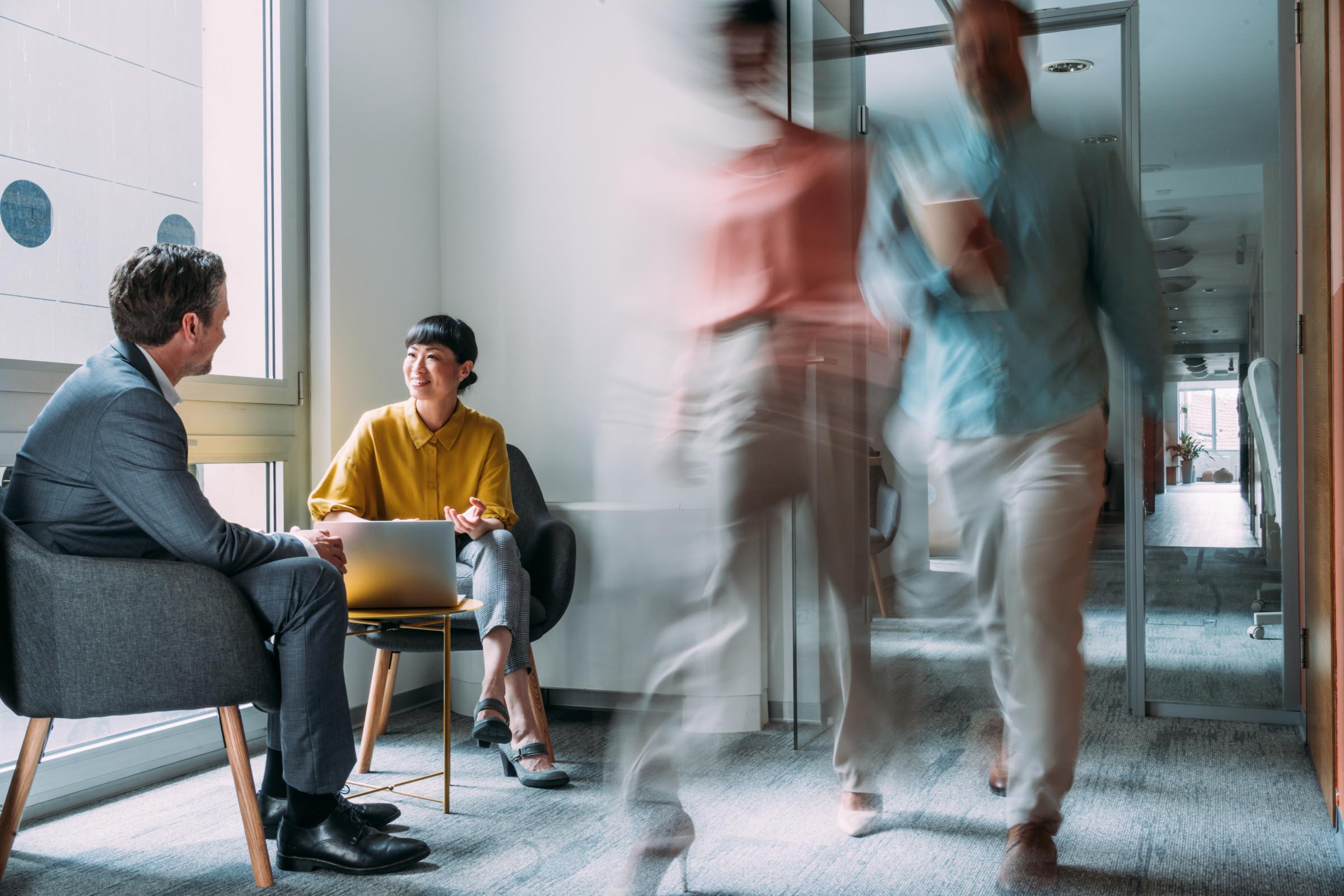 Zwei Personen sitzen und unterhalten sich in einem Büroflur, die Frau mit einem Laptop. Unscharfe Figuren suggerieren Bewegung und vermitteln eine geschäftige, dynamische Atmosphäre.