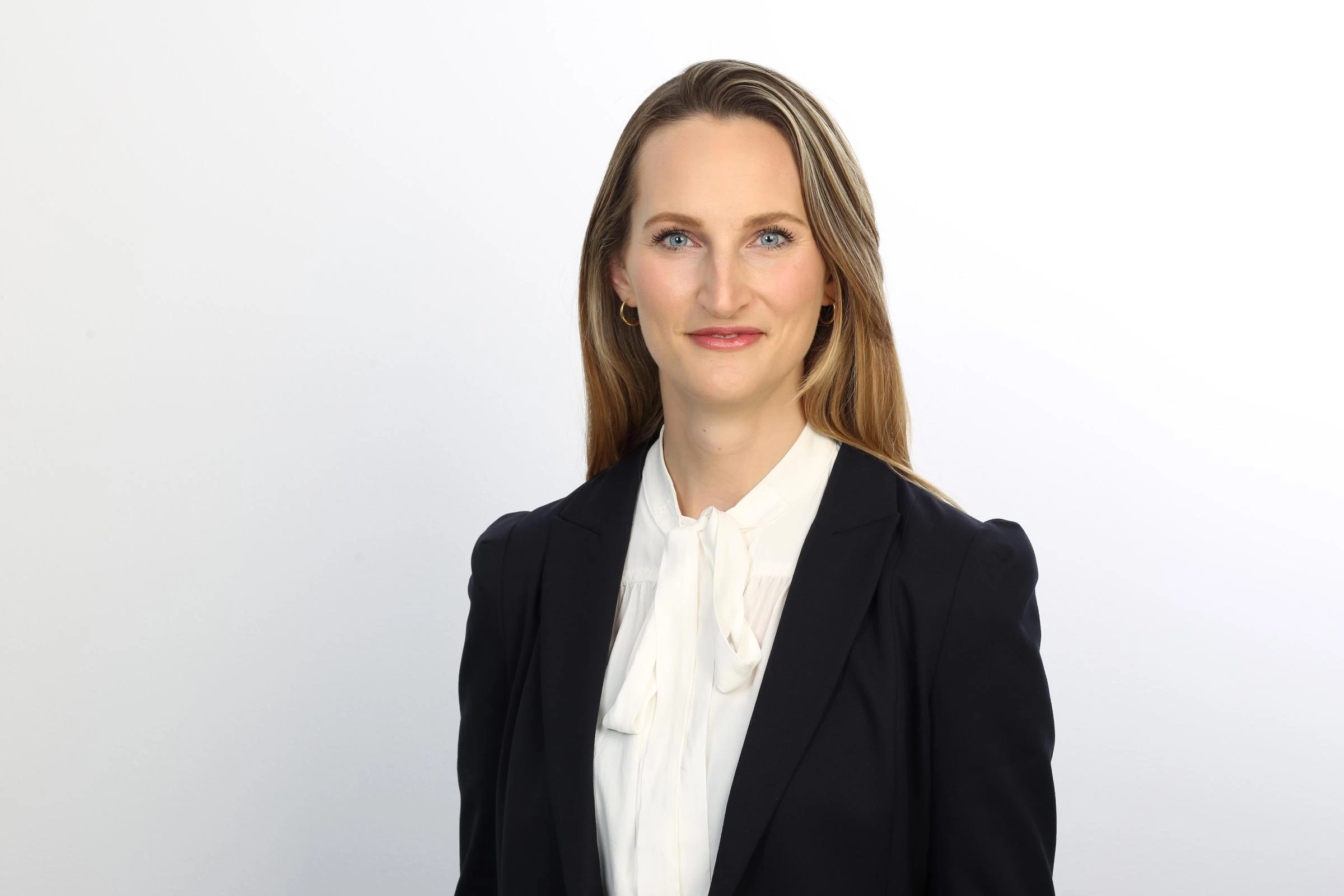 Eine junge Frau mit lange, blonden Haaren, mit weißer Bluse und schwarzem Jackett schaut leicht lächelnd in die Kamera.