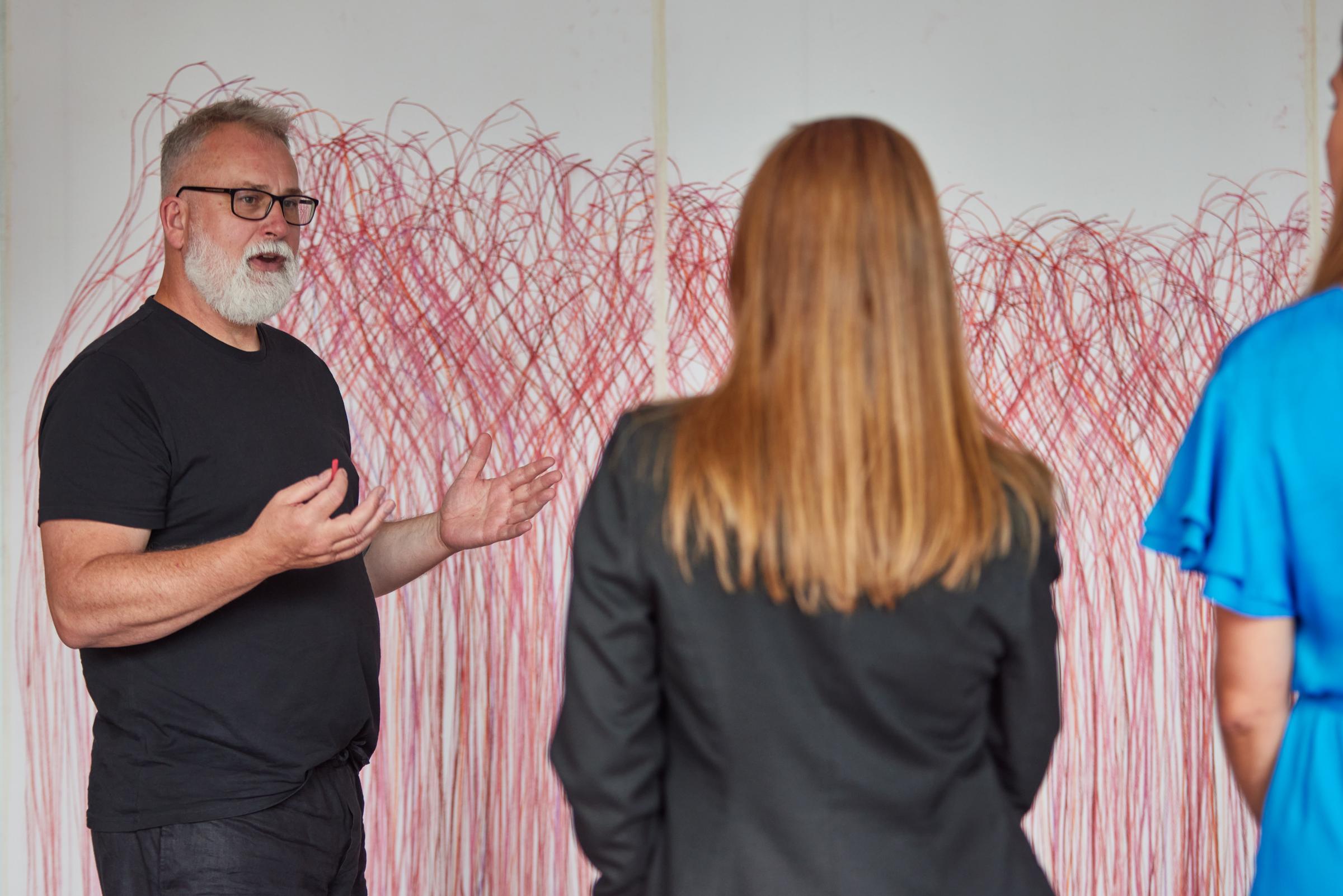 Vor einem mit roten Kreidefarben gemalten großen Wandbild steht ein Mann, der zwei anderen Menschen das Bild erläutert.