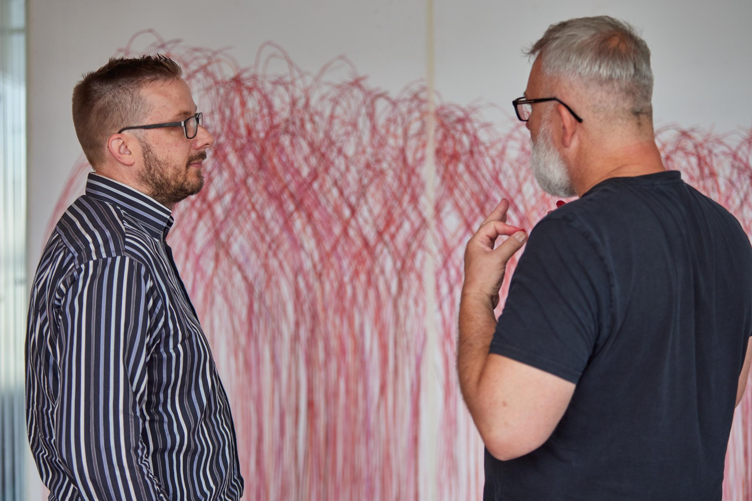 Vor einem mit roten Kreidefarben gemalten großen Wandbild stehen zwei Männer, die über das Bild sprechen.