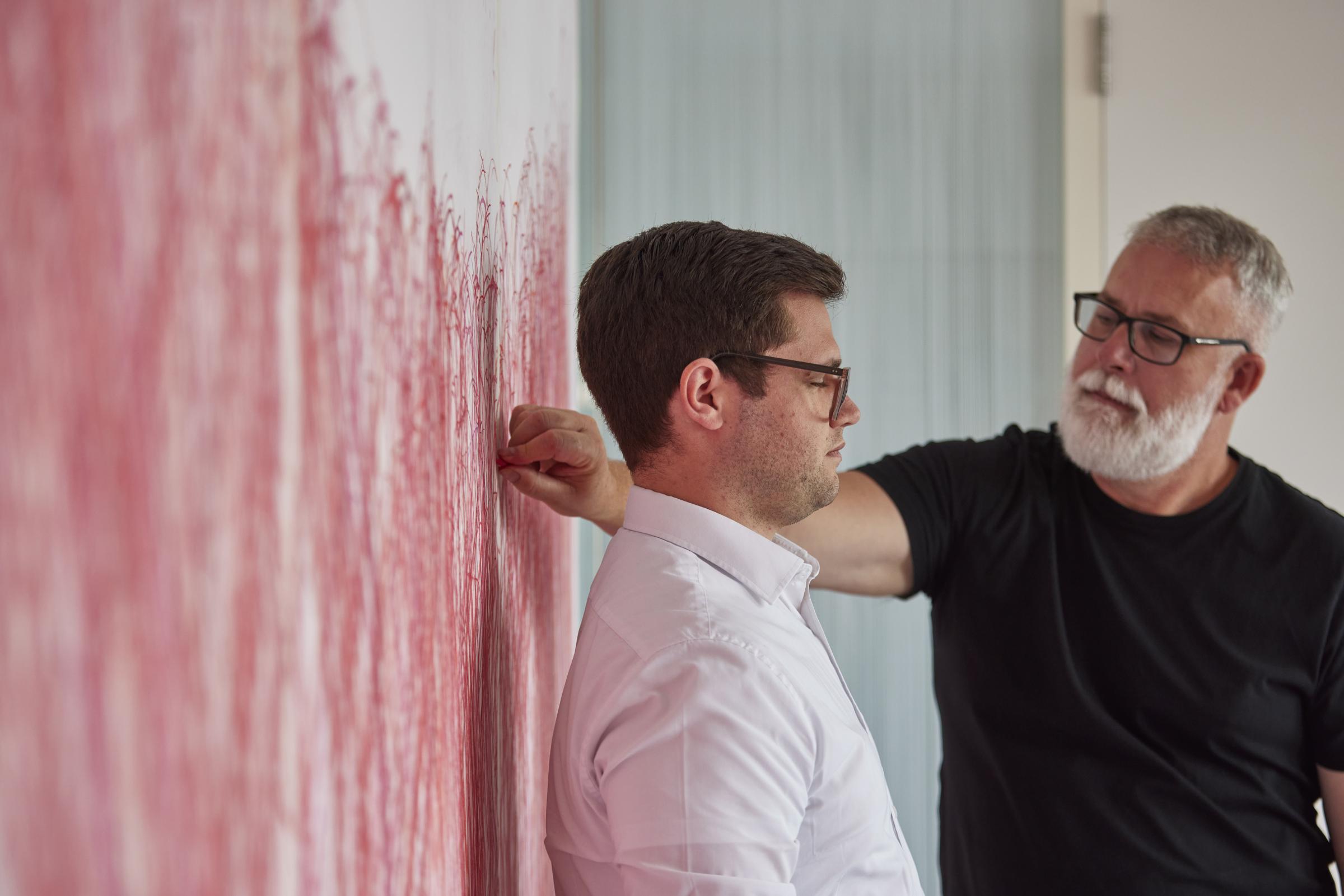 Ein Mann zeichnet den Umriss eines anderen Mannes mit roter Kreide auf ein großes Wandbild, vor dem der andere Mann mit dem Rücken steht.