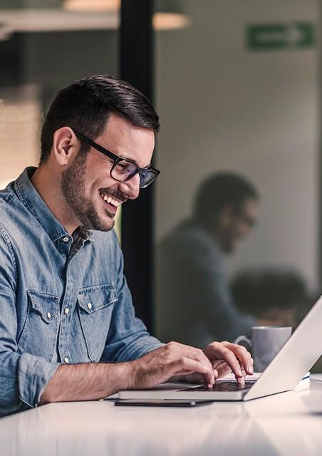 Ein junger Mann mit Brille und Jeanshemd arbeitet lächelnd am Laptop
