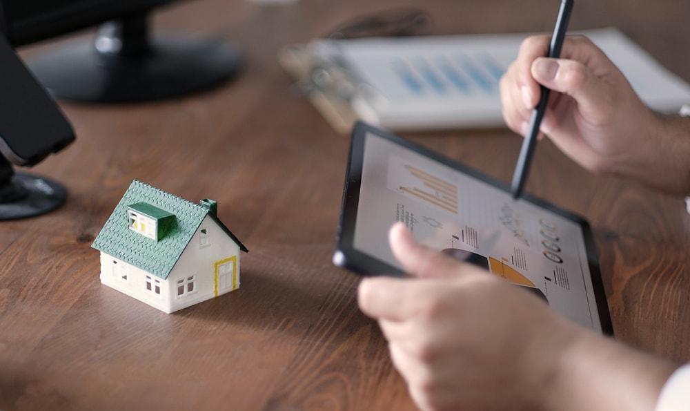 Eine Person arbeitet mit einem Stift auf einem Tablet, auf dem Diagramme angezeigt werden, daneben steht ein kleines Modellhaus auf einem Holztisch. Die Atmosphäre ist konzentriert und analytisch.