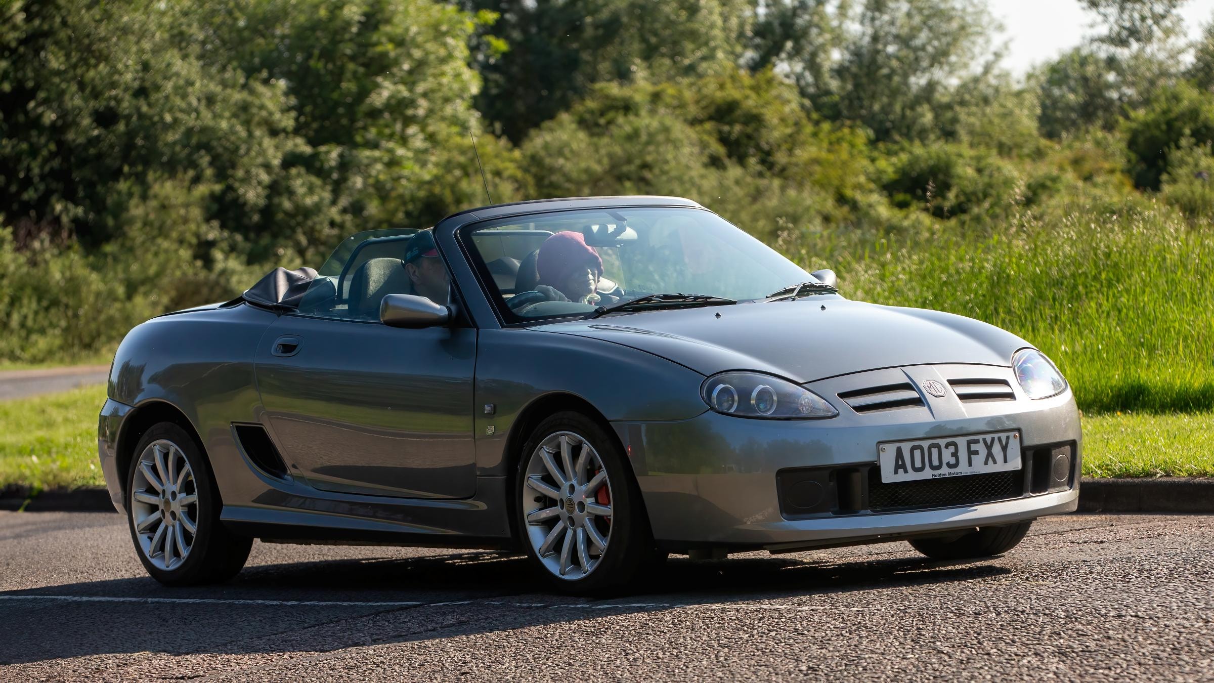 Ein silberner MG F Oldtimer fährt auf einer leeren Straße. Im Hintergrund sind grüne Bäume und Wiesen sichtbar.