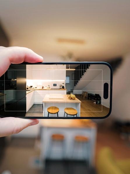 Eine Hand hält ein Smartphone und nimmt eine moderne Küche auf. Das Telefon zeigt eine weiße Insel mit zwei Hockern, die im Kontrast zu dem verschwommenen Raum im Hintergrund steht.