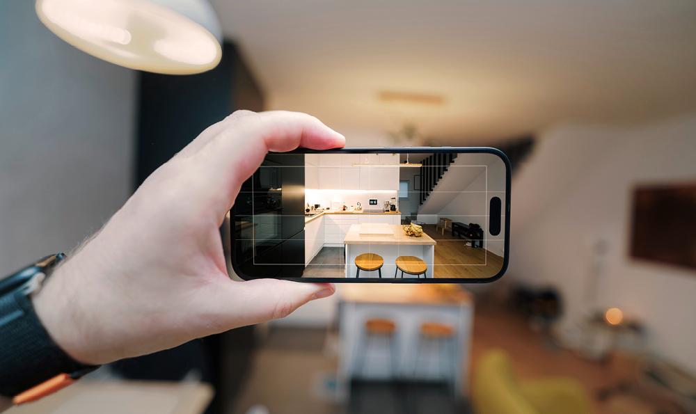 Eine Hand hält ein Smartphone, das eine stilvolle Küche mit eleganten schwarzen und weißen Schränken und hölzernen Barhockern erfasst. Die Szene strahlt Modernität und schickes Design aus.