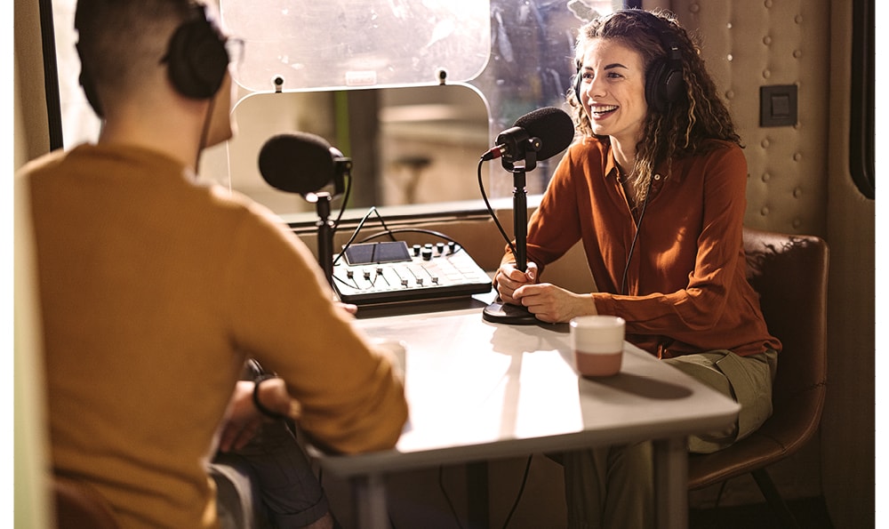 An einem Tisch sitzen ein Mann und eine Frau mit Kopfhörern, beide scheinen in ein Gespräch vertieft zu sein, das sie im Rahmen einer Podcastaufzeichnung über Micros aufnehmen.