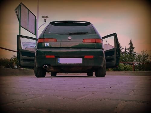 Alfa Romeo 145 mit grüner Karosserie in Heckansicht auf einem Parkplatz vor einem Wald, Türen sind offen